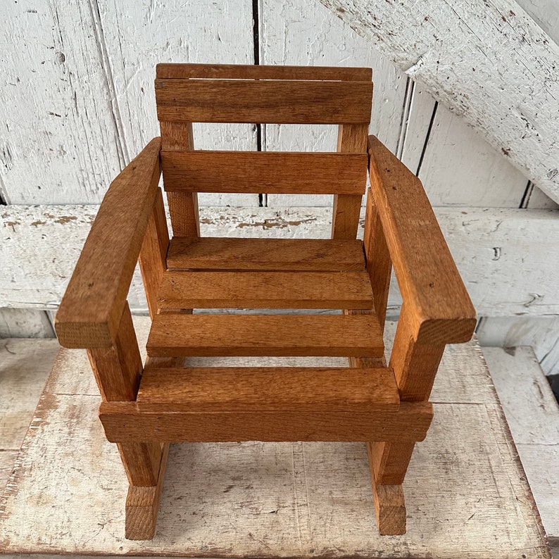 Peut inclure: Une petite chaise Adirondack en bois avec une finition naturelle. La chaise a une assise et un dossier &agrave; lattes, avec des accoudoirs. Le bois a une teinte brun-rouge&acirc;tre chaude. La chaise est con&ccedil;ue &agrave; des fins d&eacute;coratives.