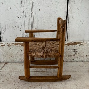 May include: A miniature wooden rocking chair with a woven seat. The chair has a natural wood finish and a traditional, rustic design. The seat is made of woven straw or a similar material, and the chair has rockers.