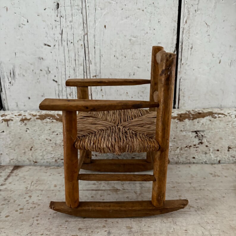 May include: A miniature wooden rocking chair with a woven seat. The chair has a natural wood finish and a traditional, rustic design. The seat is made of woven straw or a similar material, and the chair has rockers.