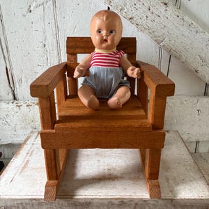 Peut inclure: Une poup&eacute;e vintage est assise dans un petit rocking-chair en bois. La poup&eacute;e porte un haut ray&eacute; rouge et blanc et un short &agrave; rayures bleues et blanches. La chaise est en bois teint&eacute; et a un design classique.