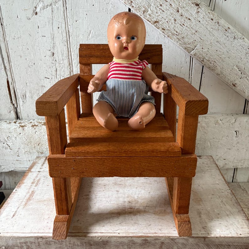 Peut inclure: Une poup&eacute;e vintage est assise dans un petit rocking-chair en bois. La poup&eacute;e porte un haut ray&eacute; rouge et blanc et un short &agrave; rayures bleues et blanches. La chaise est en bois teint&eacute; et a un design classique.