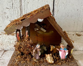 Pode incluir: Um presépio de madeira vintage com um telhado coberto de musgo. A cena inclui figuras de Maria, José, o menino Jesus e animais. As figuras estão dentro de um estábulo rústico de madeira.
