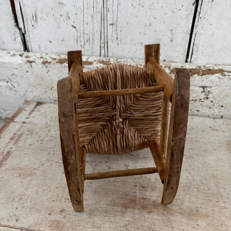 May include: A miniature wooden rocking chair with a woven straw seat. The chair is made of light brown wood and has a classic design. The seat is square and woven with a natural fibre. The chair is a miniature size.