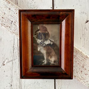 May include: A framed sepia-toned photograph of a dog wearing a hat and bow tie, with a cigarette in its mouth. The frame is made of dark brown wood. The dog is the focal point of the image.