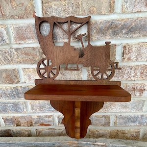 May include: A wooden shelf with a vintage car design. The shelf is made of dark brown wood and features a detailed cutout of an antique car with spoked wheels. The shelf is mounted on a decorative bracket, perfect for displaying small items.