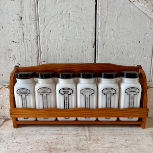 May include: A wooden spice rack holding six white glass spice jars with black lids. Each jar is labeled with a different spice name in black text, including "Garlic Salt" and "Cinnamon". The rack is set against a white wooden background.
