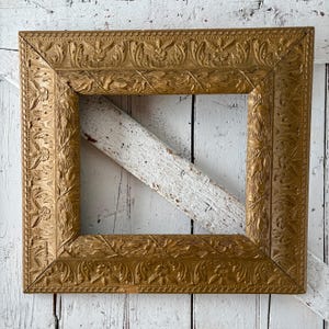 May include: Ornate gold-colored picture frame with detailed floral and leaf carvings. The frame is square and has a weathered appearance. A white wooden support is visible behind the frame.