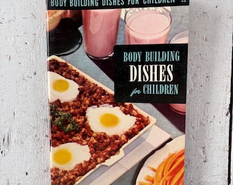 May include: A vintage cookbook titled "Body Building Dishes for Children" with a retro cover design. The cover features images of meals, including eggs, ground meat, and pink smoothies. The book is displayed on a distressed white wooden surface.