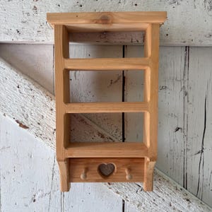 May include: A light brown wooden wall shelf featuring three open storage tiers and a bottom section with two small pegs and a central heart-shaped cutout. The unit is constructed from natural wood with visible grain patterns and a smooth finish.