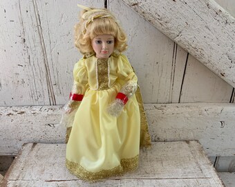 Puede incluir: Muñeca de porcelana con cabello rubio rizado, vestida con un vestido amarillo pálido con adornos dorados y detalles rojos. La muñeca tiene una expresión serena y está colocada sobre un fondo de madera blanca desgastada.