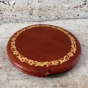 Op de afbeelding: Een ronde, roodbruine leren doos met een gouden bloemenrand. De doos heeft een metalen sluiting en vertoont tekenen van slijtage, wat wijst op de vintage aard ervan. Het ontwerp is symmetrisch en elegant.