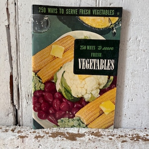 May include: A vintage cookbook cover titled "250 Ways to Serve Fresh Vegetables." The cover features a colorful illustration of cooked corn on the cob, cauliflower, red beans, and other vegetables. A dark green label displays the title.