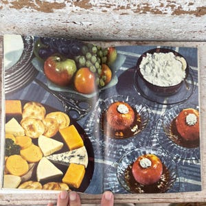 May include: A vintage magazine spread featuring a cheese and cracker platter, a bowl of cottage cheese, and baked apples in glass dishes. Grapes and apples are in a silver dish. The scene is set on a blue and white striped tablecloth.