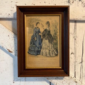 Puede incluir: Grabado antiguo enmarcado que representa a dos mujeres con vestidos de época, ambientado en un jardín. El grabado está en un marco de madera oscura con un borde interior dorado. El texto "LA CORBEILLE" es visible en la parte inferior.