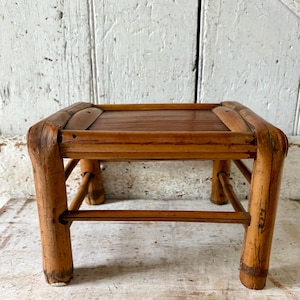 May include: A small, square bamboo stool with a slatted top and four sturdy legs. The natural bamboo is a warm, light brown color, and the stool appears to be vintage or antique. The design is simple and functional, suitable for various uses.