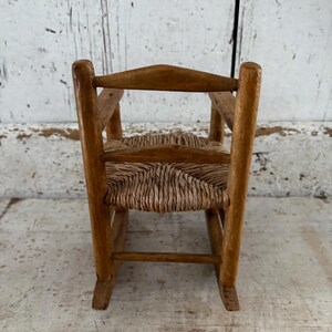 May include: A miniature wooden rocking chair with a woven seat. The chair has a natural wood finish and a traditional design. The seat is made of woven straw or a similar material. A charming piece of vintage furniture.