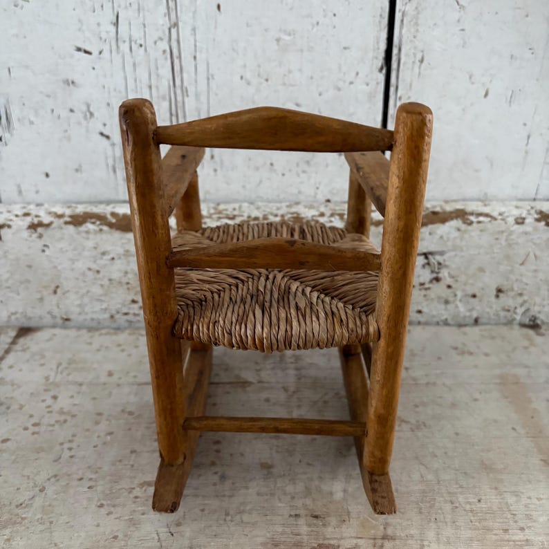 May include: A miniature wooden rocking chair with a woven seat. The chair has a natural wood finish and a traditional design. The seat is made of woven straw or a similar material. A charming piece of vintage furniture.