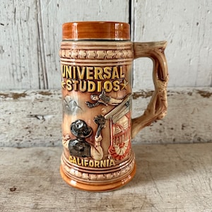 May include: A tan ceramic beer stein featuring a relief design of Universal Studios California. The mug displays a figure in a hat, a building structure, and the text Universal Studios California. It has a textured handle and a smooth rim.