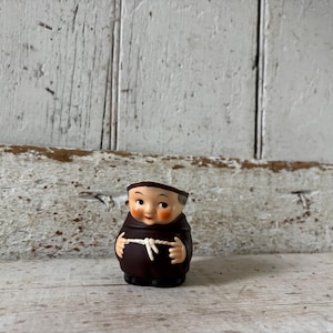 Puede incluir: Pequeña figura de cerámica vintage de un monje. La figura viste una túnica marrón con un cinturón de cuerda blanco. El monje tiene una cara sonriente con mejillas rosadas y una gorra negra. El fondo es una superficie de madera blanca desgastada.