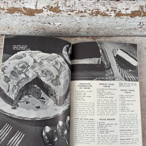 May include: An open cookbook page featuring recipes for desserts like Chocolate Marshmallow Ice Cream and Apricot Angel Cream. A slice of cake with meringue topping is visible, along with serving utensils.