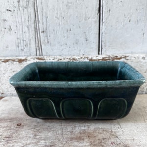 May include: A rectangular, dark teal ceramic dish with a glossy finish. The dish has a slightly textured rim and three curved indentations on the front. The interior is a darker shade of teal, and the dish sits on a weathered wooden surface.