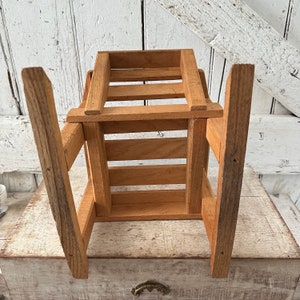 Peut inclure: Petite caisse en bois avec une finition naturelle. La caisse est de forme rectangulaire avec des lattes verticales sur les c&ocirc;t&eacute;s et un dessus &agrave; lattes. Les pieds sont longs et inclin&eacute;s, ce qui lui donne un aspect robuste. Le grain du bois est visible.
