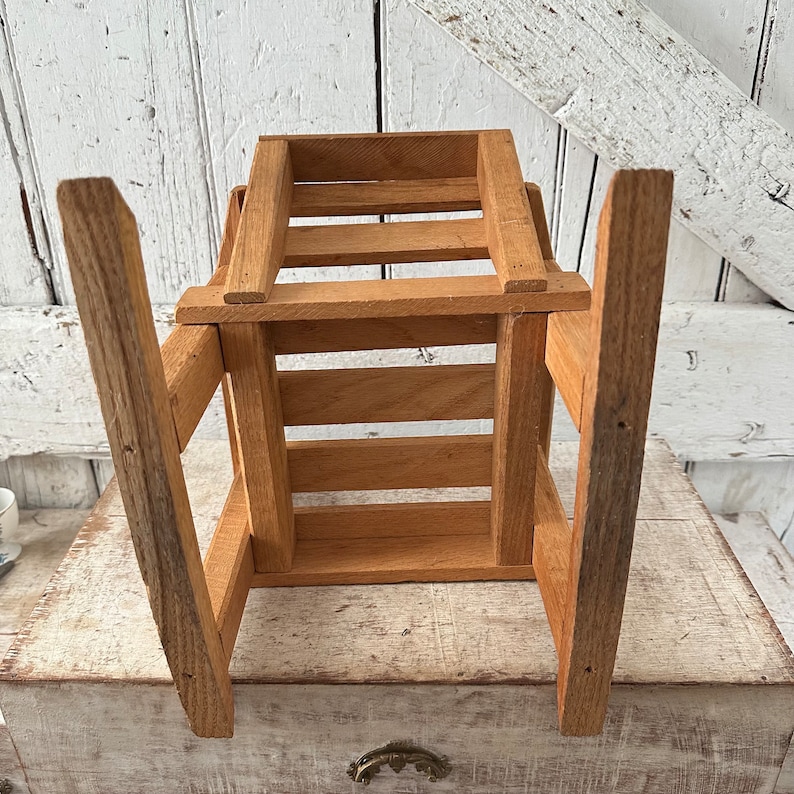 Peut inclure: Petite caisse en bois avec une finition naturelle. La caisse est de forme rectangulaire avec des lattes verticales sur les c&ocirc;t&eacute;s et un dessus &agrave; lattes. Les pieds sont longs et inclin&eacute;s, ce qui lui donne un aspect robuste. Le grain du bois est visible.