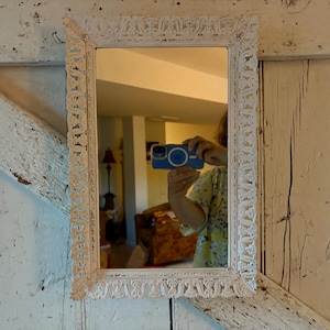 Pode incluir: Um espelho retangular branco e ornamentado com uma moldura decorativa. A moldura apresenta padrões intrincados e repetitivos. O espelho reflete uma sala com uma pessoa segurando um telefone azul. O espelho é montado em uma superfície branca e de madeira.