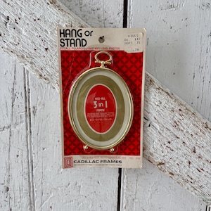 May include: A vintage gold-toned oval picture frame with a red and white backing. The frame is labelled "HANG or STAND" and "FITZALL POLAROID - INSTANT-LOAD PHOTOS." The frame is 8.89 x 11.43 cm and can be used as a 3-in-1 frame.