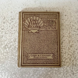 May include: A vintage book titled "Cyr's First Reader" with a brown cover and intricate design. The book is published by Ginn & Company, Publishers.
