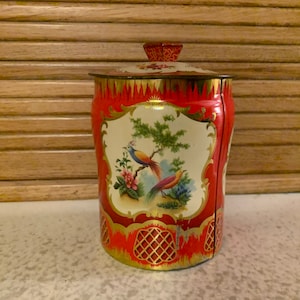 May include: A vintage red tin canister with a lid, featuring a decorative bird and floral design on a white background. The canister has gold trim and a lattice pattern at the base, showcasing a classic, ornate style.