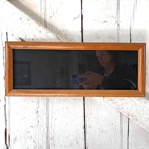 May include: Rectangular wooden framed mirror. The frame is a light brown color. The mirror's surface is dark and reflective, showing a person holding a phone. The mirror is mounted on a white, weathered wooden surface.