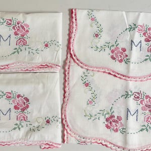 May include: Four white rectangular linens featuring cross-stitched pink rose floral wreaths and the letter M embroidered in blue. Each piece is finished with a scalloped edge trimmed in pink thread, creating a matching set of decorative textiles.