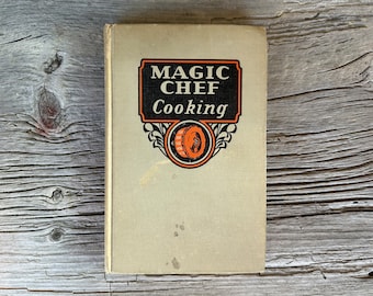 May include: A vintage cookbook titled "MAGIC CHEF Cooking" with a cream-colored cover. The title is in black and white, with an orange and black emblem. The book is slightly worn, with some stains on the cover, and rests on a weathered wooden surface.