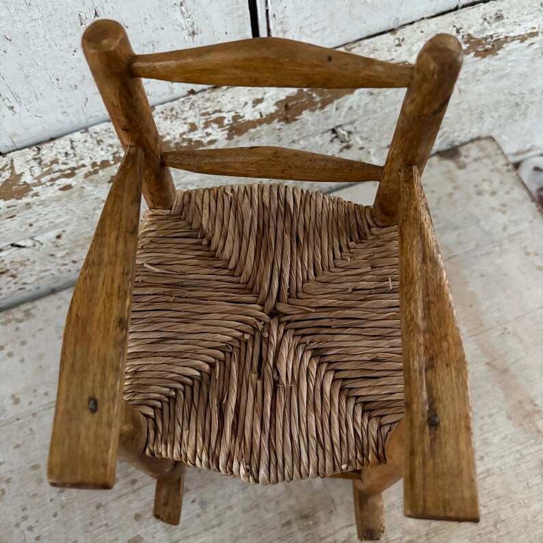 May include: Small wooden chair with a woven straw seat and a light brown finish. The chair has a classic design with a slatted back and four legs. The seat is made of tightly woven straw, providing a rustic and natural look. The chair is suitable for a doll or small child.