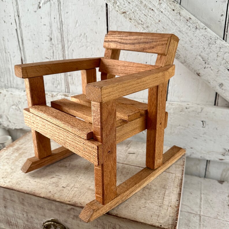 Peut inclure: Petite chaise &agrave; bascule en bois naturel. Elle est compos&eacute;e d'une assise &agrave; lattes, d'accoudoirs et d'un dossier, tous fabriqu&eacute;s &agrave; partir de pi&egrave;ces de bois rectangulaires. Les patins de la chaise sont visibles, sugg&eacute;rant un design classique et artisanal.