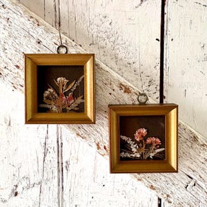 May include: Two square wall frames with metallic gold borders and dark brown backings. Each frame contains a pressed dried flower arrangement with white, pink, and green tones. Each frame features a small circular metal loop at the top for hanging.