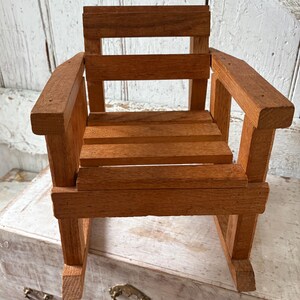 Peut inclure: Petite chaise &agrave; bascule en bois naturel. Le si&egrave;ge et le dossier sont &agrave; lattes, avec des accoudoirs. Le grain du bois est visible, et la chaise semble faite &agrave; la main. Con&ccedil;ue pour une poup&eacute;e ou un petit jouet.
