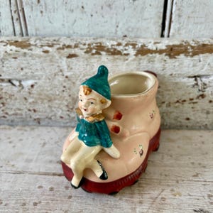 Könnte beinhalten: Vintage Keramik-Blumentopf in Stiefelform, bemalt in Rosa- und Rottönen. Eine kleine Figur eines Elfen mit tealfarbener Mütze und Jacke sitzt auf dem Stiefel. Der Stiefel hat florale Akzente und ist wahrscheinlich ein dekorativer Artikel.