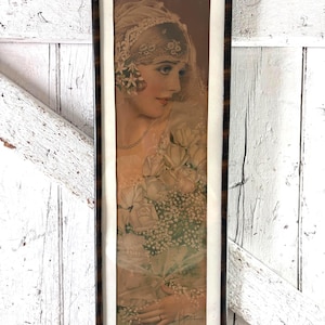 Puede incluir: Una impresión vertical enmarcada que muestra el retrato de una persona con un velo de encaje y tocado, sosteniendo un gran ramo de rosas blancas y paniculata. La obra está dentro de un marco de madera marrón oscuro con un borde blanco fino.