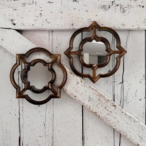May include: Two wall mirrors featuring dark bronze-toned frames with a quatrefoil-inspired geometric shape. Each mirror has a central reflective glass pane surrounded by a decorative, symmetrical frame with curved edges and pointed corners.