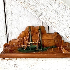 May include: Wooden relief carving depicting a coastal scene with a two-masted sailboat and buildings. The boat is painted green, and the buildings and landscape are a natural wood tone. The carving is mounted on a wooden base.