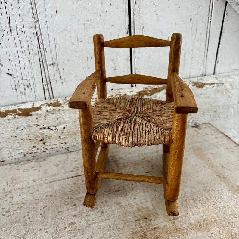 May include: A miniature wooden rocking chair with a woven straw seat. The chair has a natural wood finish and armrests. The small chair is ideal for a doll or decorative display. The chair's dimensions are approximately 30 cm in height.