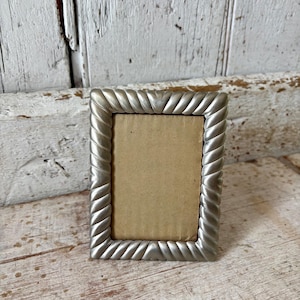 May include: A rectangular picture frame with a metallic silver finish and a prominent twisted rope-style border. The frame features a plain, light tan cardboard insert centered within the metallic frame structure.