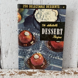 May include: A vintage cookbook titled "250 Delectable Desserts" with a cover featuring images of baked apples and a bowl of whipped cream. The cover has a black banner with the words "Dessert Recipes". The book is on a wooden surface.