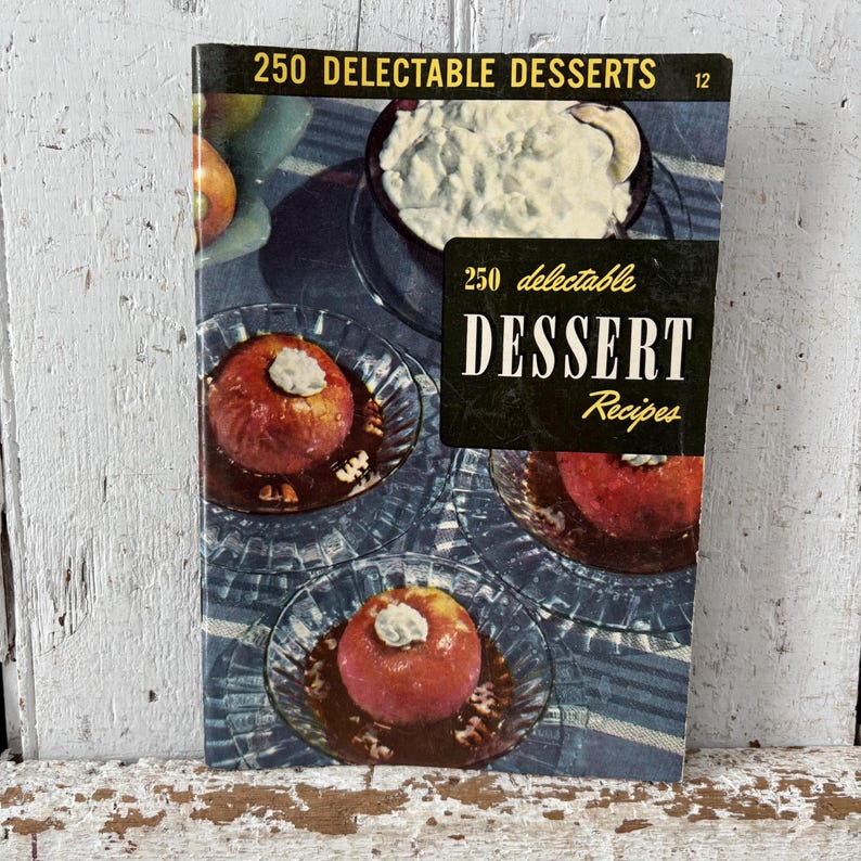 May include: A vintage cookbook titled "250 Delectable Desserts" with a cover featuring images of baked apples and a bowl of whipped cream. The cover has a black banner with the words "Dessert Recipes". The book is on a wooden surface.