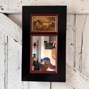 Peut inclure: Miroir rectangulaire à cadre noir. La partie supérieure présente une impression encadrée de moutons dans un champ. La partie inférieure est un miroir reflétant une pièce. Le cadre est rouge foncé.