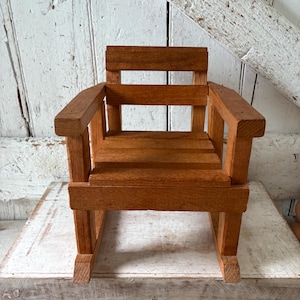 Peut inclure: Petite chaise en bois naturel. Le dossier et l'assise sont &agrave; lattes, avec des accoudoirs. Le grain du bois est visible. La chaise semble &ecirc;tre faite pour un enfant ou une poup&eacute;e. La chaise est en bois.