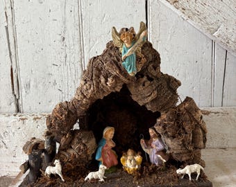 Pode incluir: Um presépio em miniatura com figuras de Maria, José, o menino Jesus e um anjo. As figuras estão dentro de uma estrutura rústica em forma de casca com pequenas ovelhas brancas. O anjo segura um pergaminho.