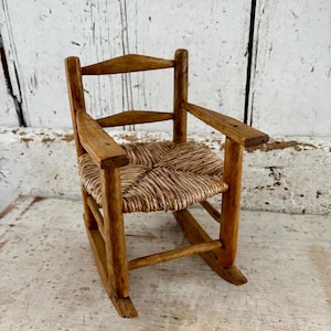May include: A small, antique wooden rocking chair with a woven straw seat and armrests. The chair has a natural wood finish and is designed for a doll or small child. The chair is approximately 30 cm tall.